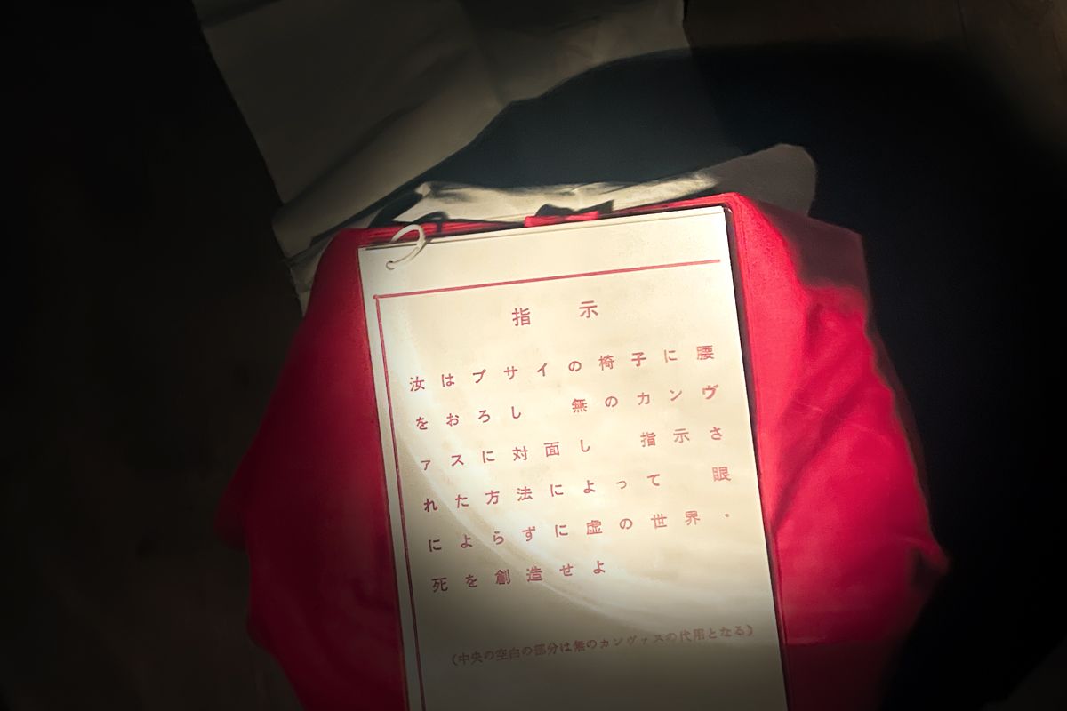 《9の無のカンヴァスと9のプサイの椅子と9の超未来的方法による松澤宥展V.1010》（1967）展示風景。メイン会場の母屋から蔵に移動すると、入口で大型の携帯電灯を手渡された。携帯電灯で暗闇を照らしながら1階の展示《ヴァニシングス（消滅事）》を経て2階に上がると本作品の展示があった。まっさらなカンヴァスの前には、写真の “指示”と椅子が置かれていた。「指示　汝はプサイの椅子に腰をおろし　無のカンヴァスに対面し　指示された方法によって　眼によらずに虚の世界・死を創造せよ」